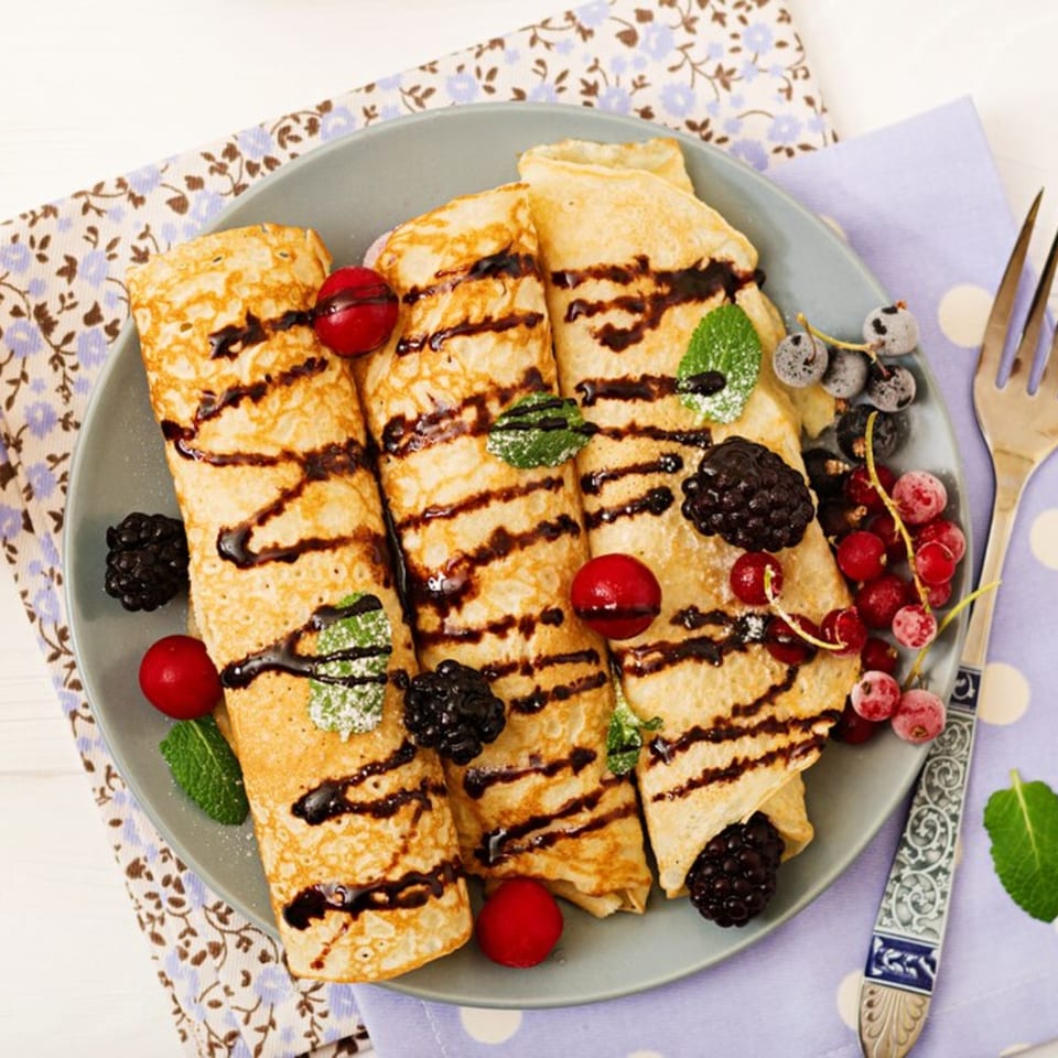 Three chocolate-drizzled crepes with fresh berries and mint on a gray plate.